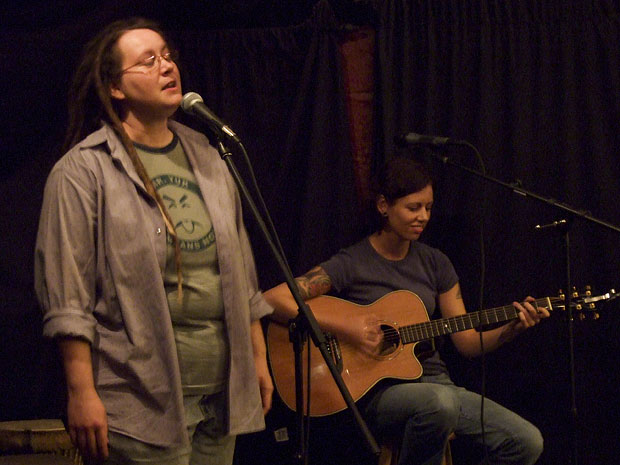 A photo of two women. The one closest to the camera is singing into a mic. The one further away on the right side of the image is sitting down playing guitar.