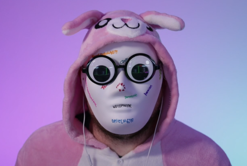 A person in a pink bunny suit on a light background gradient from purple to light blue. They are wearing a solid white mask with sporadic hand-writing on it in different colors. They're wearing larger googly eye glasses with a pair of white headphone wires visible coming out from behind the mask