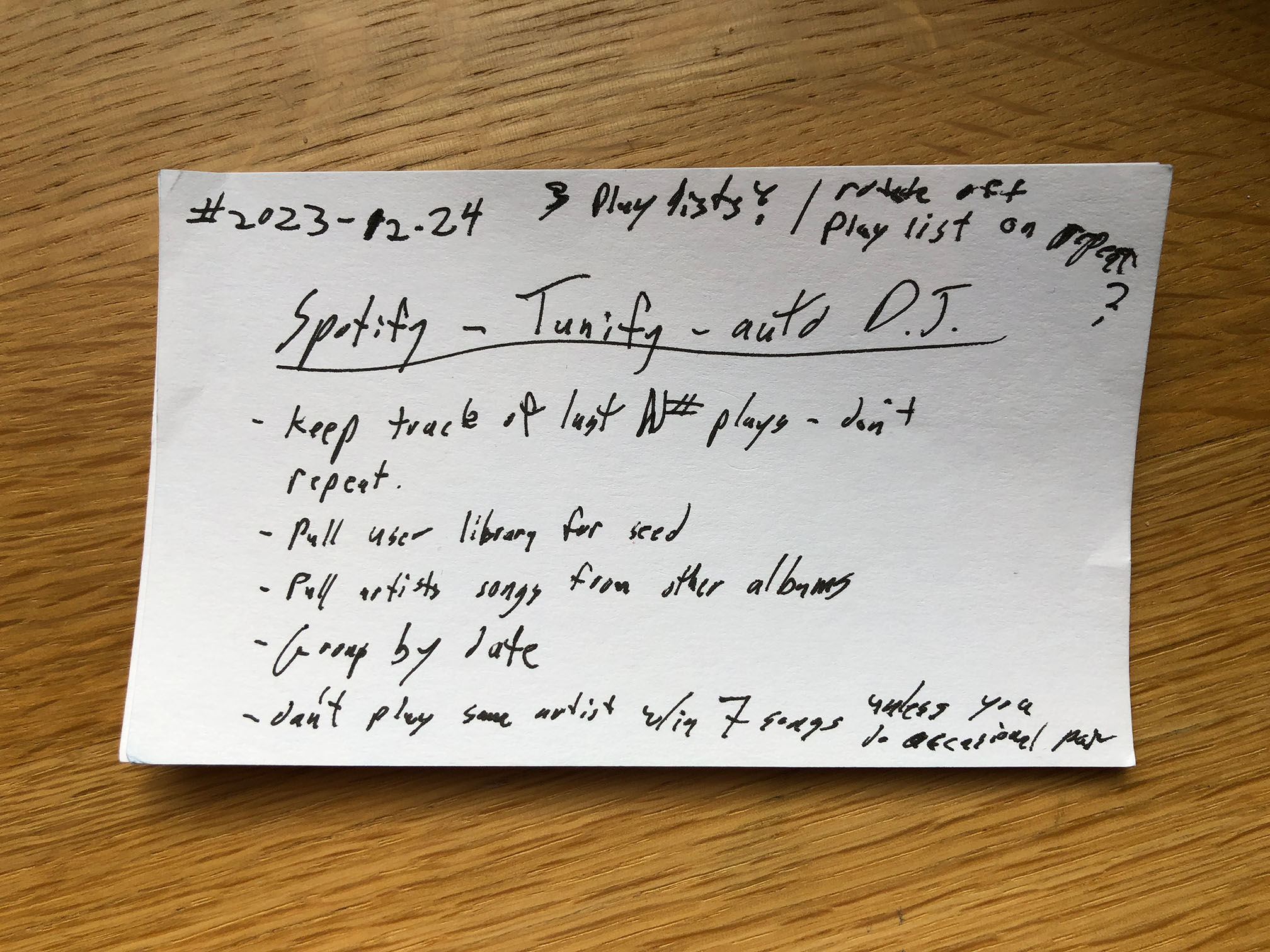 A photo of a 3x5 note card laying on a wood surface. The underlined title of the card is 'Spotify ~ Tunify ~ auto D.J.' Under the title is a list with the following items: 1 keep track of the last N# of plays. don't repeat. 2 pull user library for seed. 3 pull artist songs from other albums. 4 group by date. 5 don't play the same artist within 7 songs unless you do an occasional pair. Above the title is the date 2023 ~ 12 ~ 24 and 3 playlists? and roll off playlist on repeat?
