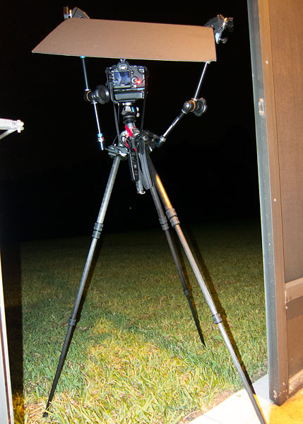 A camera sitting on a tripod. The camera is facing away. There are two flexible clamp amrs holding a piece of cardboard above the camera to keep it dry