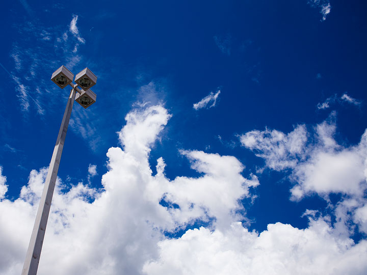 Bright white clouds against a clear, deep blue sky with a lamppost extending up from the lower left portion of the frame. The clouds fill the bottom half of the frame with the sky visible in the upper half. The line between them is a ragged movement up and down from the shape of the clouds. The light post is a square post made of a sun-baked gray metal. Three square boxes extend from the top of the post at ninety degree angles. Each contains a circular light.