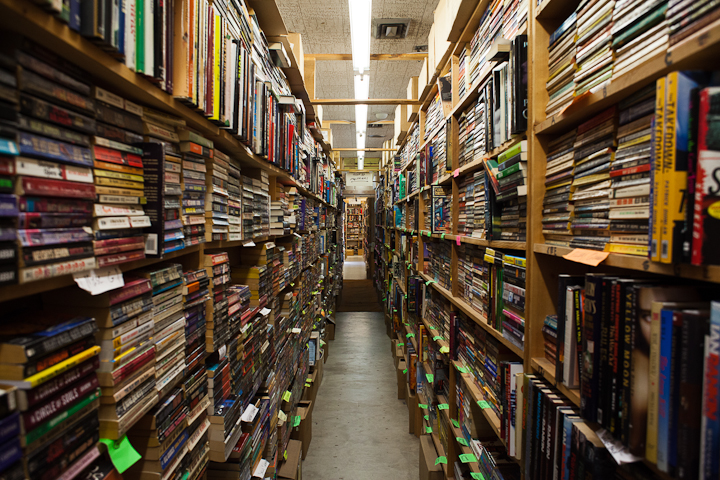 A photo looking down a narrow aisle of the Chamblin Bookmine book store. Tight wooden shelves packed full of paper-back books completely fill sides of the frame and extend to the end that's probably fifty feet away where another bookshelf faces the row. The floor is polished gray concrete with a line of flourescent lights placed directly over the aisle.