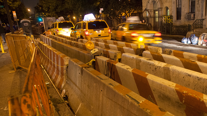 Five rows of concrete barricades sections lined up next to each other on the side of a a New York City street. Some are plain. Some have orange and white stripes. The barricades start at the lower right corner of the image and extend away from the camera toward the upper left corner. The image has an overall orange tint from the sodium vapor lights of the city. Two stopped taxi vans are in the road on the other side of the barricades along with another one showing some motion blur as it moves past.