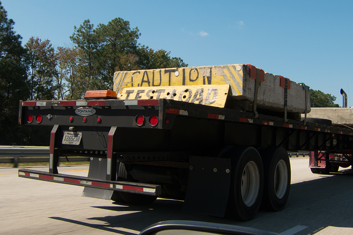 A photo of the back of a flat trailer being pulled behind a semi taken from the window of a car beside and a little behindit. The bark of the flat bed has what looks like a block of concrete on it with the words 'Caution Test Load' painted on it.