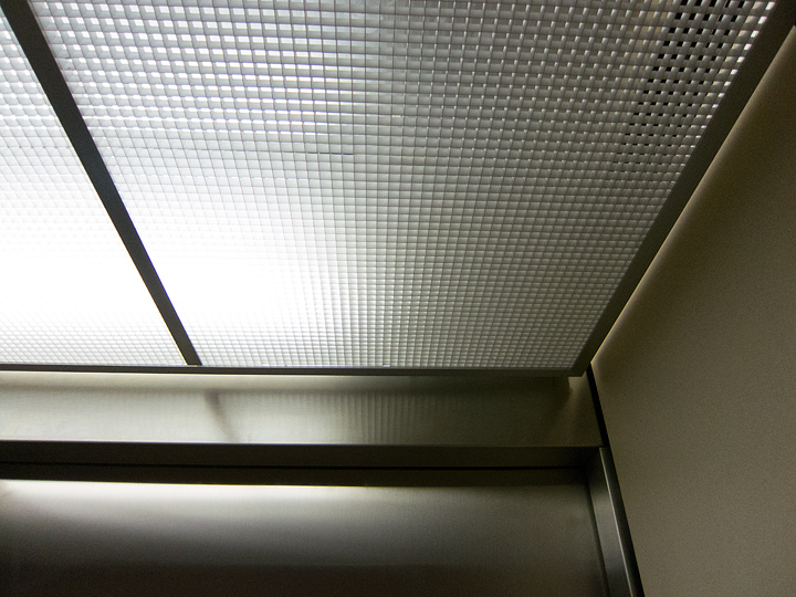 The grid over the ceiling light in an elevator. The image is taken looking at the corner from the back of the elevator zoomed in a bit. The effect is to show a portion of the grid and the upper corder of the steel door below it. The wall is visible on the right side as well. Collectively they make strong lines that compliment the grid which is made of hundreds of small squares.