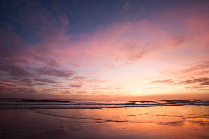 A sunrise photo over the beach. The sun isn't visible but the sky is relatively bright with golds and purples and in the clouds and blue at the top of the sky moving to a bright yellow where the sun is about to crest. The image was taken from a low angle at low tide. The sand is smooth with a thin layer of water over it reflecting the orange from the sky.
