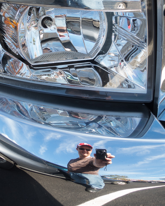 A photo taken in the reflection of a the front bumpper of a car just below the headlight. The entire image has a silver and black feel with a reflection of a blue sky with scattered clouds and myself stading up in jeans, a brown shirt, and a crimson and white alabama hat